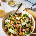 A large serving bowl of farro salad with serving utensils sticking out and some ingredients around.
