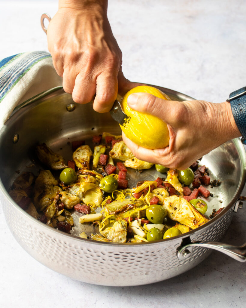 Hands zesting lemon over a hammered stainless steel saute pan with salami, artichokes and olives inside.