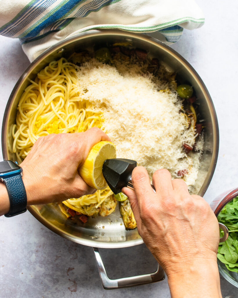 Hands juicing a lemon into a saute pan with pasta, parmesan and other ingredients inside.