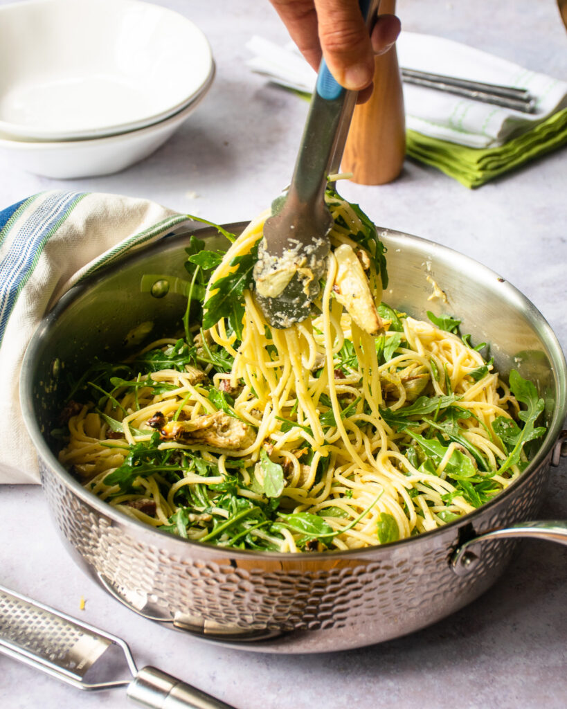 Tongs lifting pasta from a saute pan.