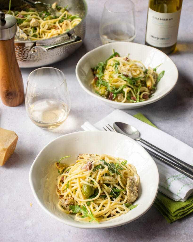 Two bowls of lemon pasta with salami, olives and artichokes on a table with a pepper mill, white wine, parmesan cheese and grater.