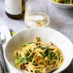 Lemon pasta with salami, olives and artichokes in a white bowl on a table with a glass of wine and a place setting.