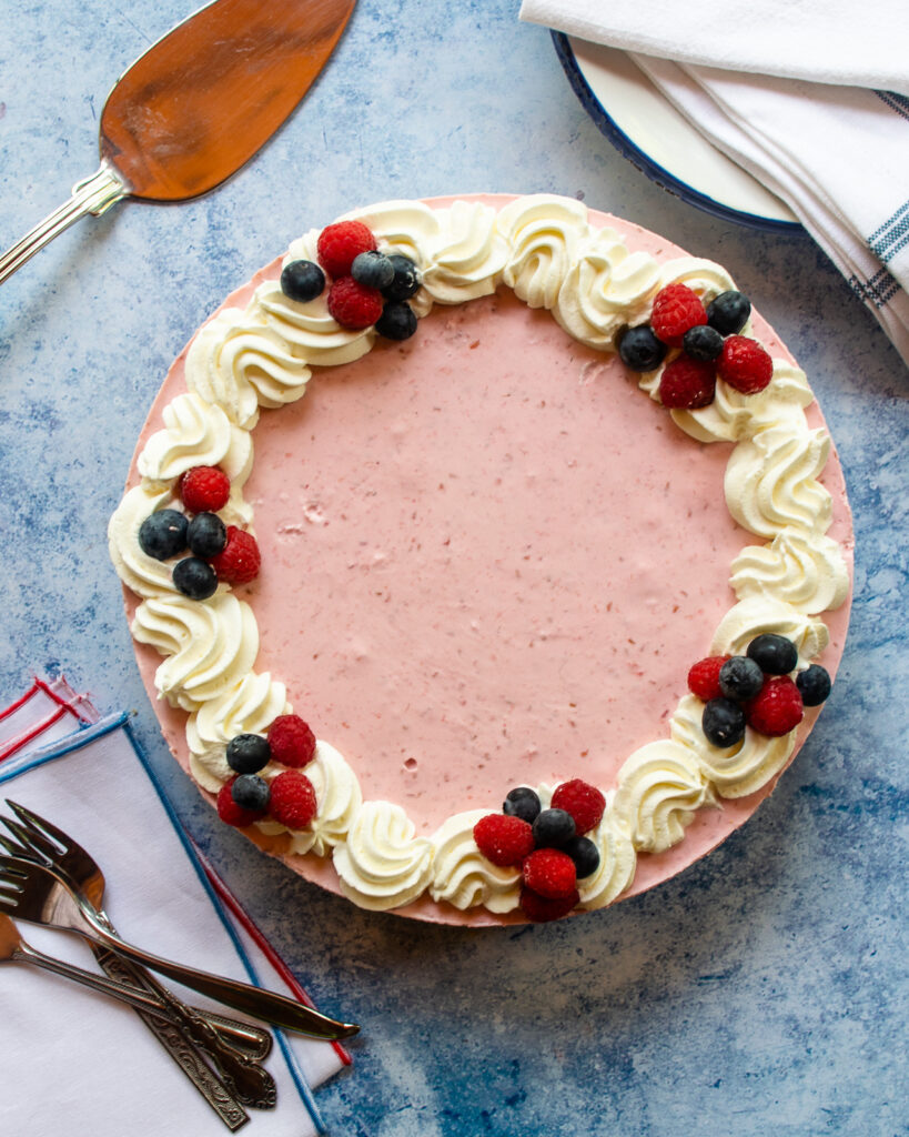 Looking straight down on a no bake cheesecake with whipped cream and berries, plates, napkins, forks and a pie server.