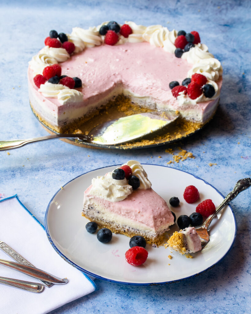 A piece of no bake layered cheesecake with a forkful removed on a blue rimmed white plate with the rest of the cheesecake in the background.