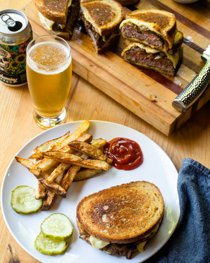 A patty melt on a white plate with fries next to a stein of beer and more patty melts on a board.