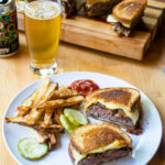 A white plate with a patty melt and some french fries, with a beer behind it and more patty melts on a wooden board.