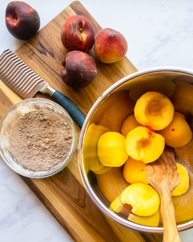 Halved peaches in a stainless steel bowl with a wooden spoon, a bowl of spice mix and some whole peaches on a wooden cutting board with a small knife.