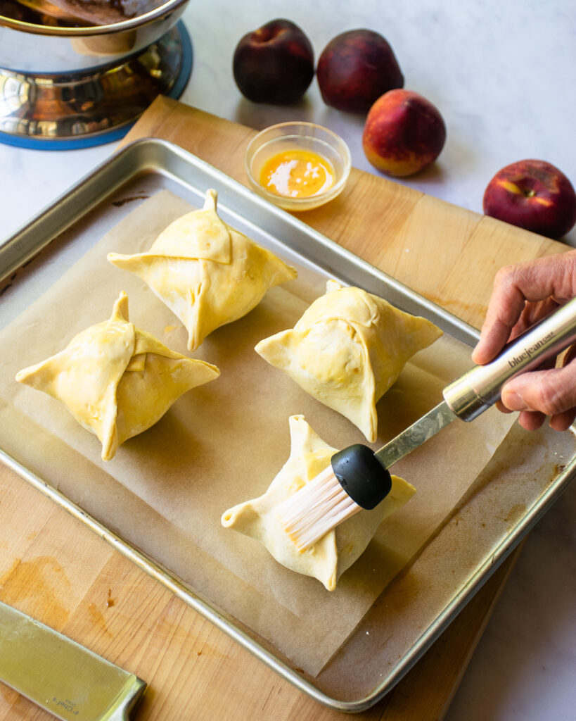 A hand brushing egg wash on peach dumplings on a baking sheet lined with parchment paper.