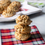 A stack of three pignoli cookies on a plaid bev nap with more pignoli cookies in the background.