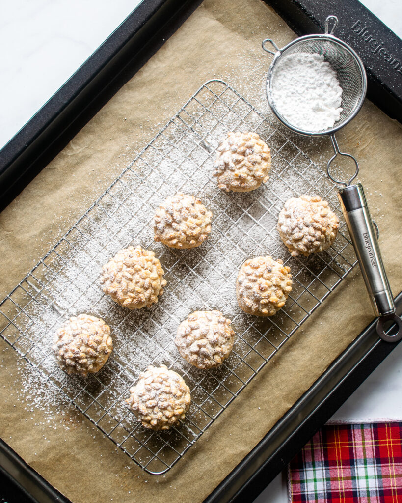 Looking down on pignoli cookies on a cooling rack, dusted with powdered sugar over a parchment paper-lined cookie sheet and a fine strainer of powdered sugar.