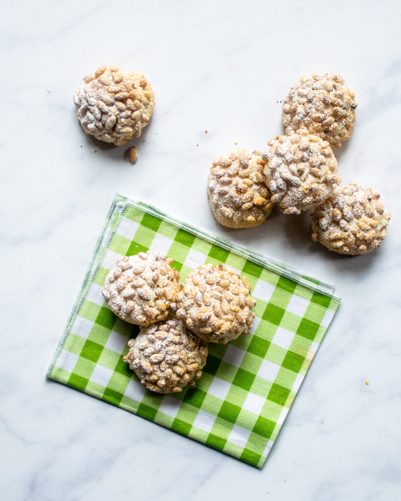 Pignoli cookies dusted with powdered sugar on a marble countertop with a few on a green checkered bev napkin.