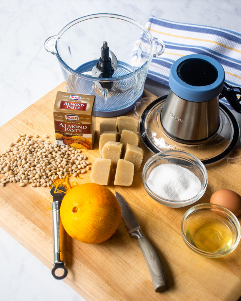 Ingredients on a cutting board - almond paste, pine nuts, an orange with a zester, an egg with a bowl of egg white, sugar, a paring knife and a mini chopper.