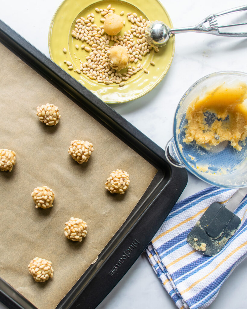 Looking down on a sheet pan partially filled with pignoli cookies next to a plate of pinenuts and a bowl of cookie dough.