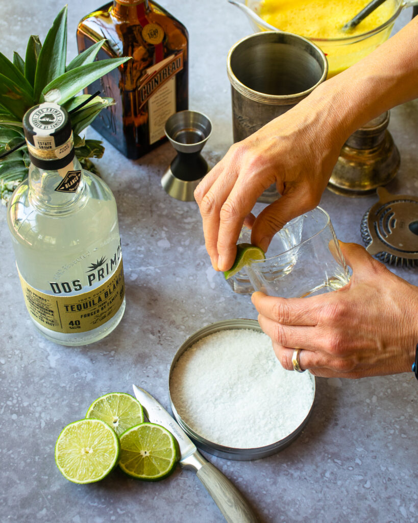 Hands rimming a rocks glass with a wedge of lime with margarita ingredients around.