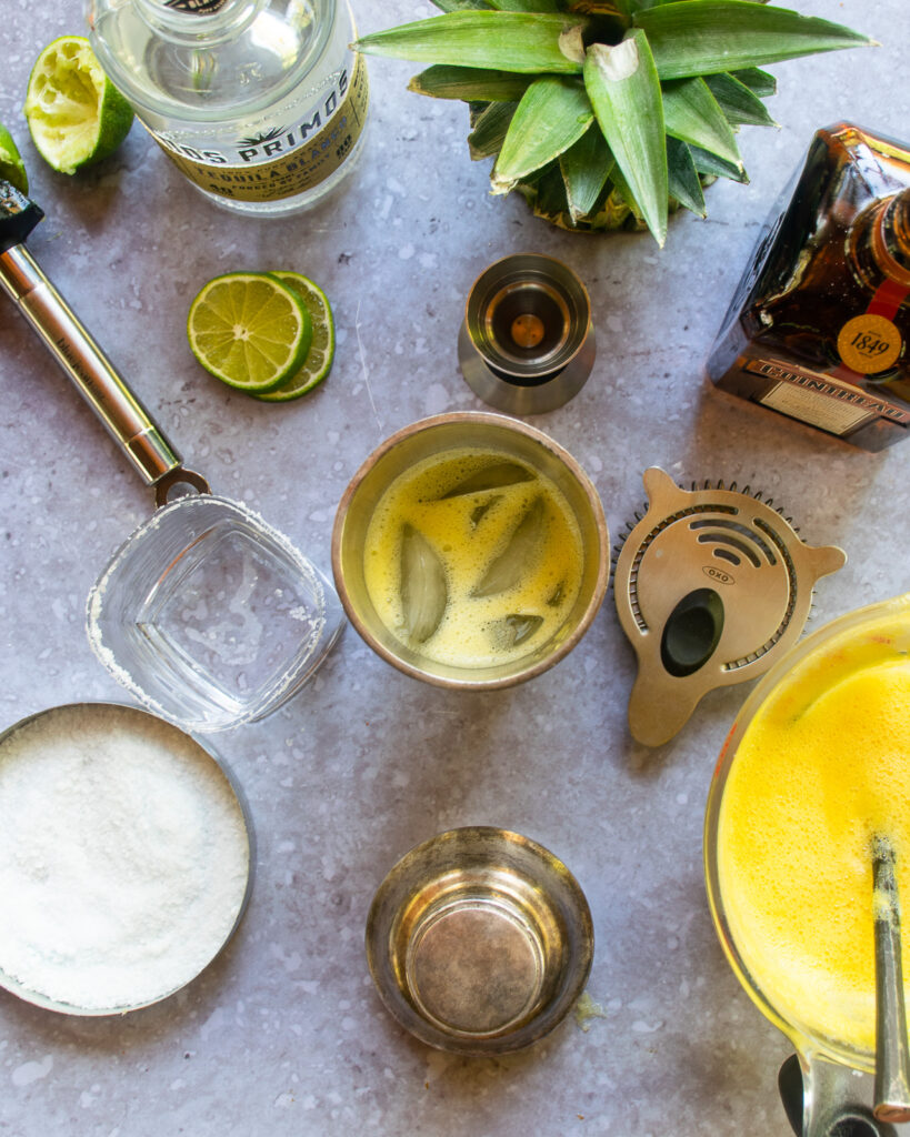 Looking down into a shaker with pineapple margarita and ingredients all around.