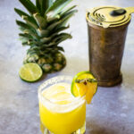 A pineapple margarita in a rocks glass with a shaker and pineapple top behind it.