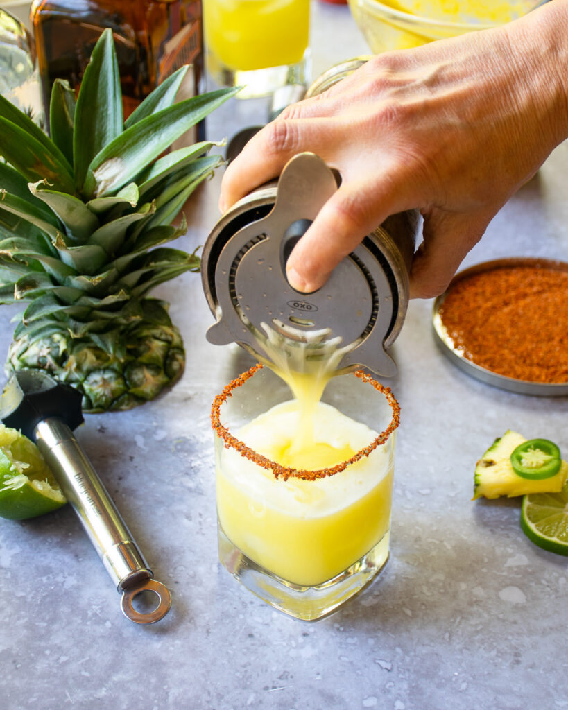 Pouring a spicy pineapple margarita out of a cocktail shaker into a Tajin rimmed rocks glass.