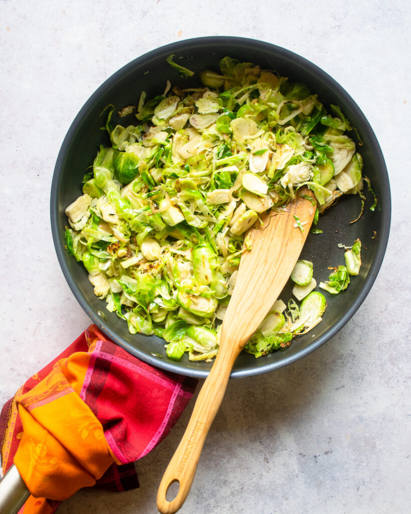 Brussels sprouts cooking in a large skillet with a wooden spoon sticking out of it.