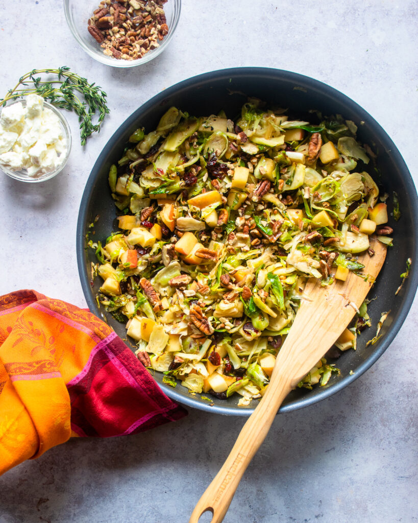 A skillet with Brussels sprouts in a large blue non-stick skillet with a wooden spoon sticking out of it and a colorful towel wrapped around the skillet handle.