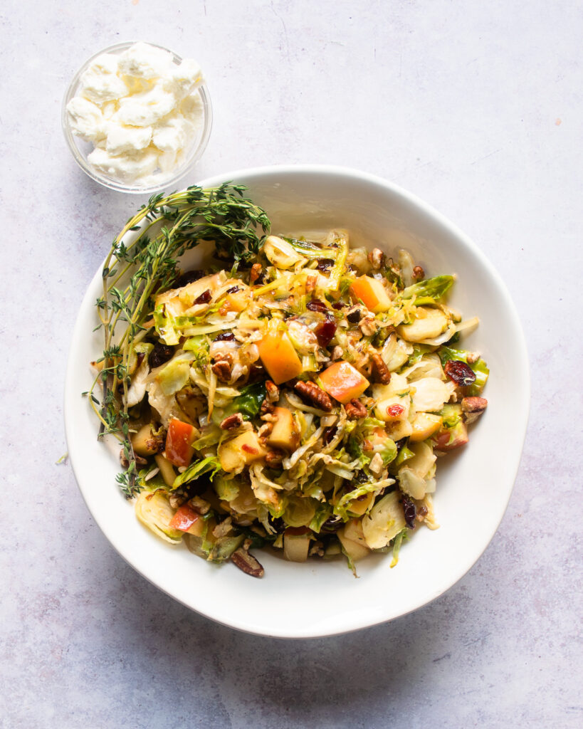 Brussels sprouts with apples and cranberries in a white bowl with a smaller bowl of goat cheese on the side.