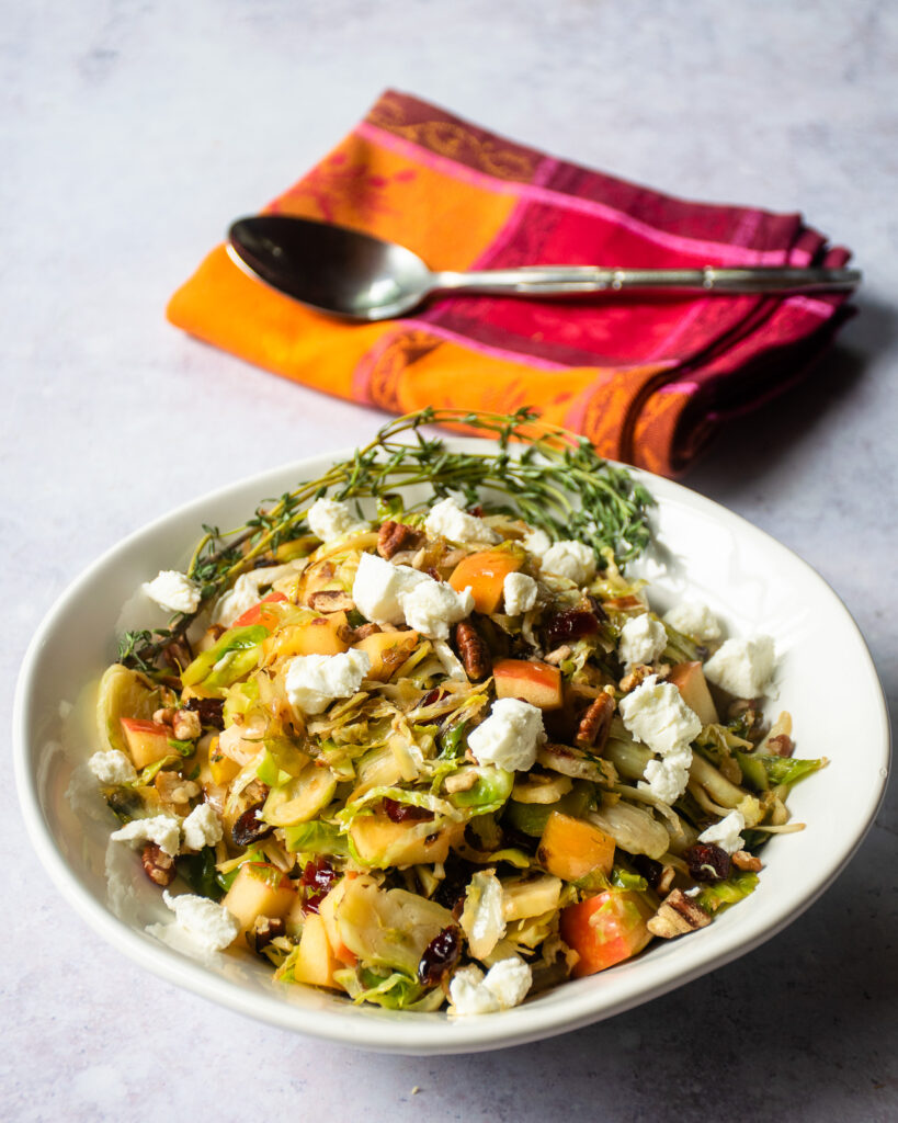 A white serving bowl of shaved Brussels sprouts with apples, cranberries and goat cheese with a colorful napkin and spoon in the background.