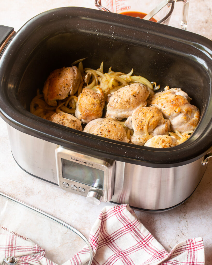 A slow cooker on a counter with browned chicken thighs and onions inside, with a red and white checked towel in front with the lid.