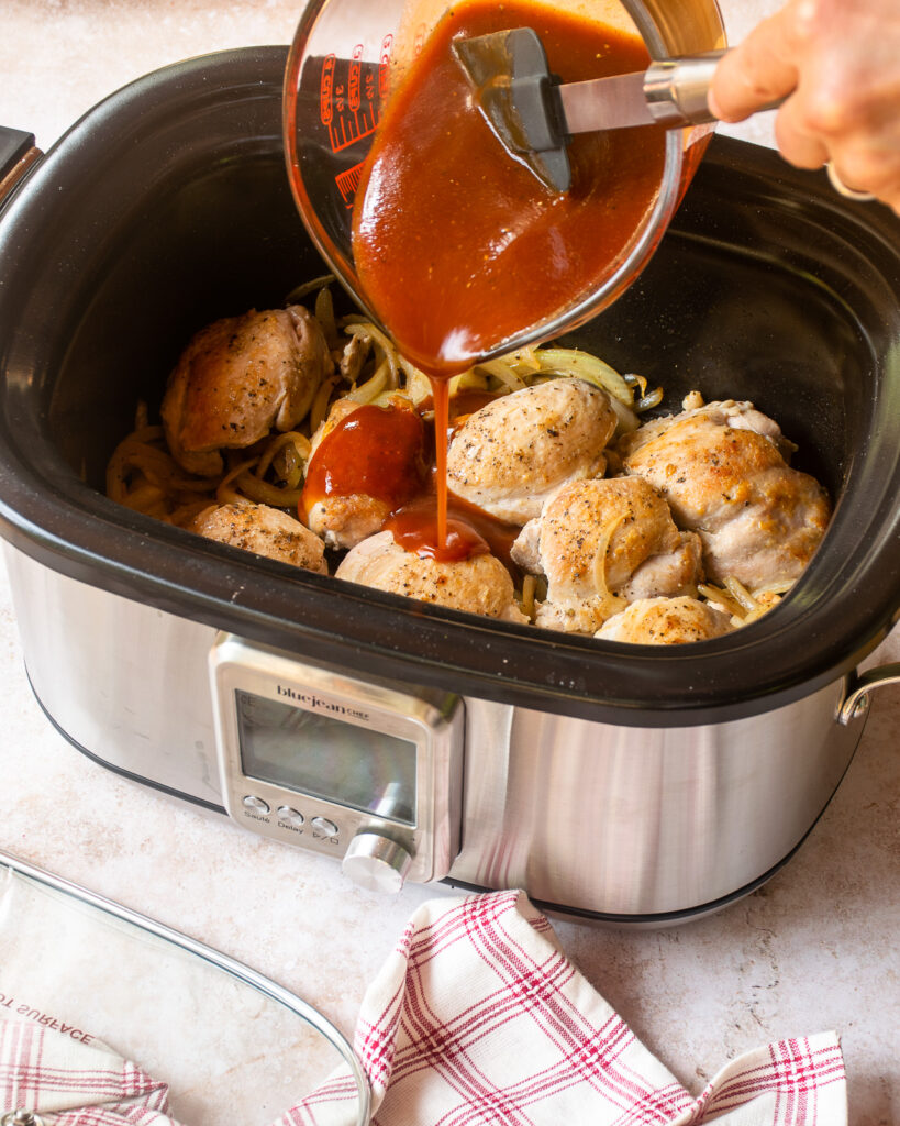 Hands pouring BBQ sauce over browned chicken thighs in a slow cooker.