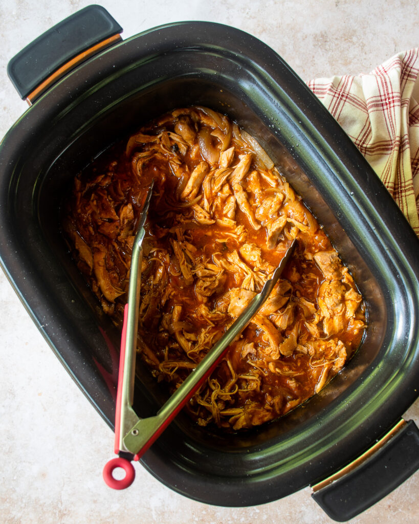 Looking straight down into a slow cooker with shredded BBQ chicken and a pair of tongs inside.