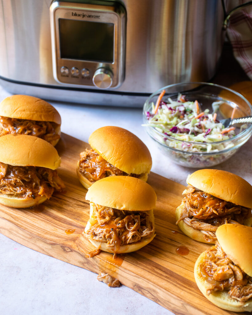 6 BBQ pulled chicken sliders on a wooden board in front of a slow cooker with a bowl of coleslaw.