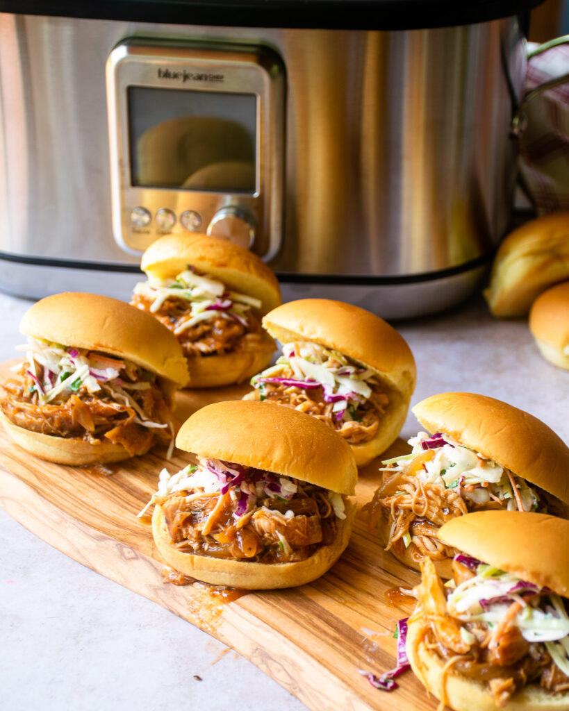 6 BBQ pulled chicken sliders on a wooden board in front of a slow cooker.