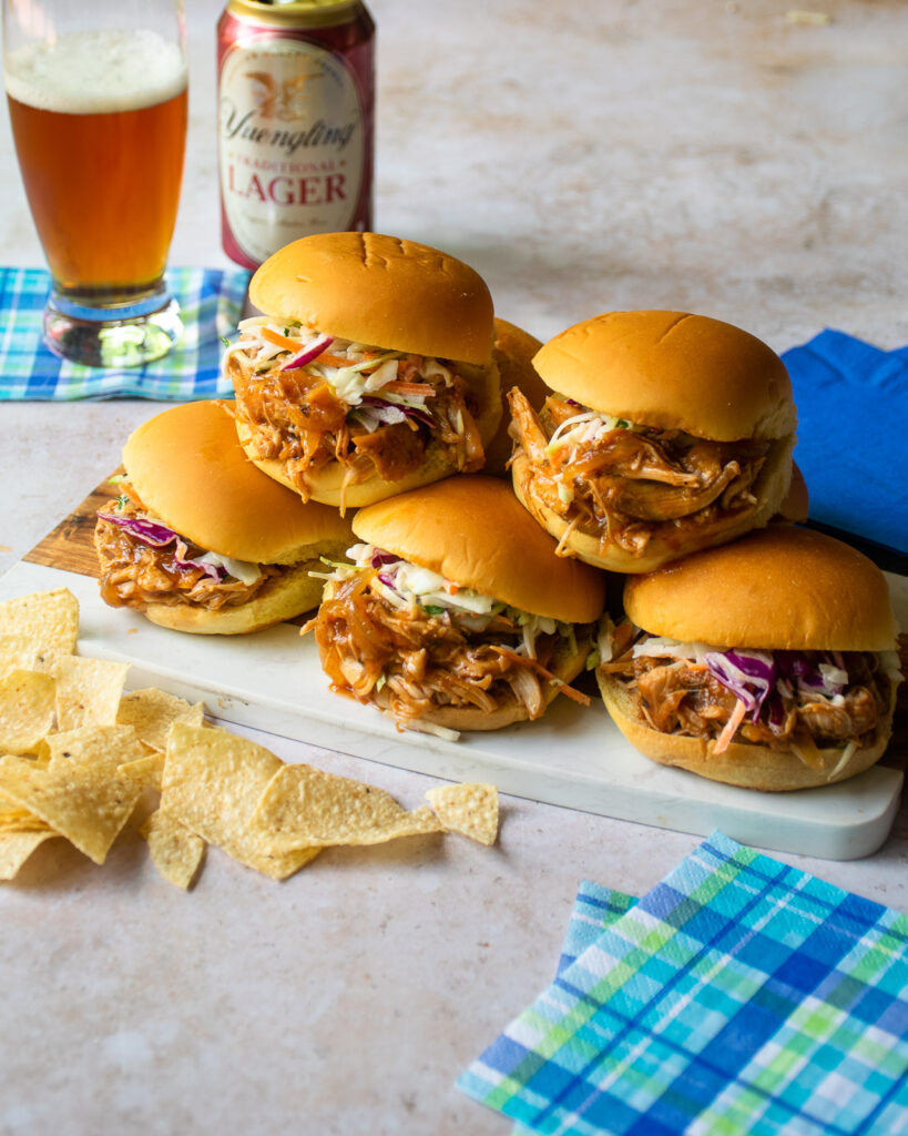 Pulled BBQ chicken sliders stacked on a little serving board with a glass and can of beer in the background and chips and bev naps in the foreground.