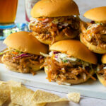 BBQ Pulled Chicken sliders stacked on top of each other with chips in the foreground and beer in the background.