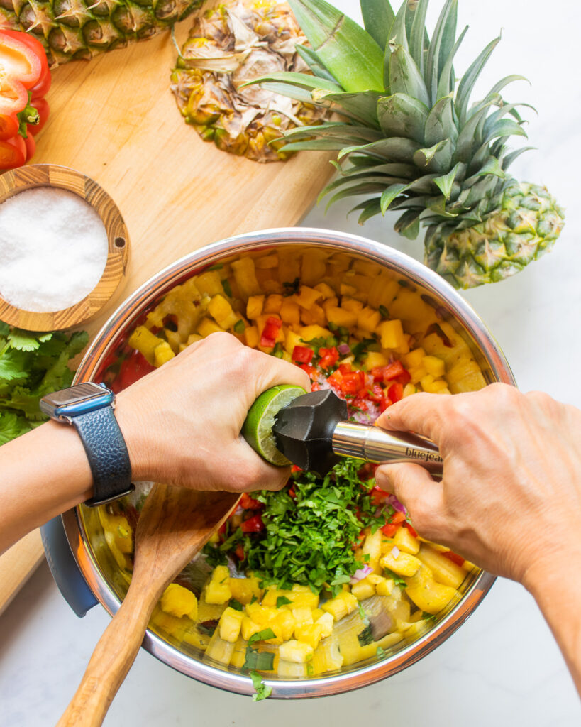 Hands squeezing lime juice into a large stainless steel bowl with pineapple salsa inside.