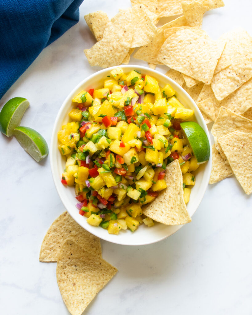 Looking straight down on a white bowl of pineapple salsa with tortilla chips, lime wedges and a blue napking.