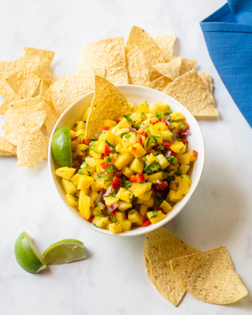 Looking straight down on a white bowl of pineapple salsa with tortilla chips, lime wedges and a blue napking.