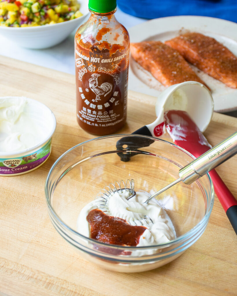 A mixing bowl on a wooden cutting board with ingredients around - sour cream, sriracha sauce, salmon and a bowl of pineapple salsa in the background.