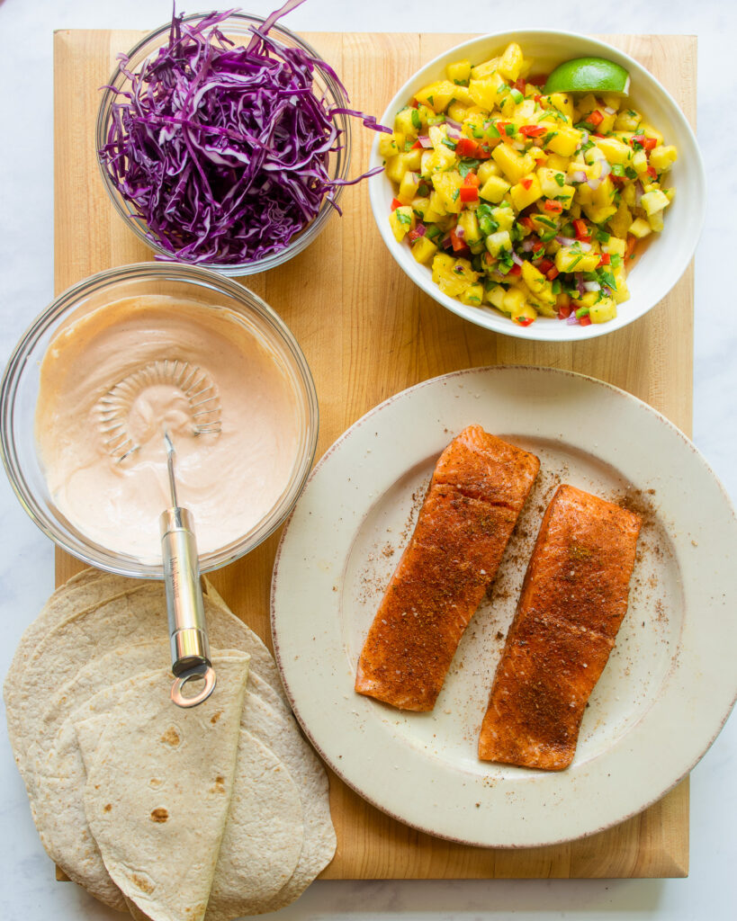 Looking down on the components of spicy salmon tacos - tortillas, seasoned salmon, spicy crema, pineapple salsa and shredded purple cabbage.