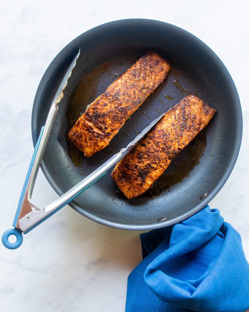 Two fillets of salmon blackened in a skillet with a pair of tongs and a blue kitchen towel.