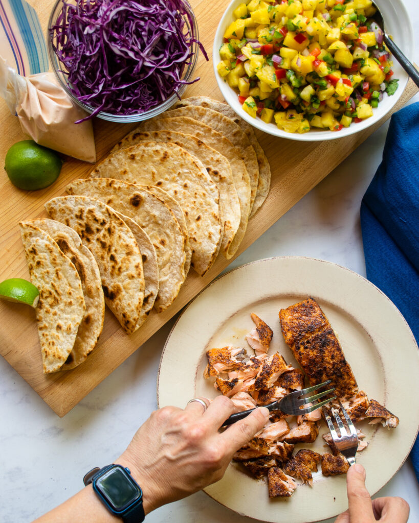 Hands flaking cooked salmon fillets on a plate next to fried tortilla shells, mango salsa and shredded red cabbage.