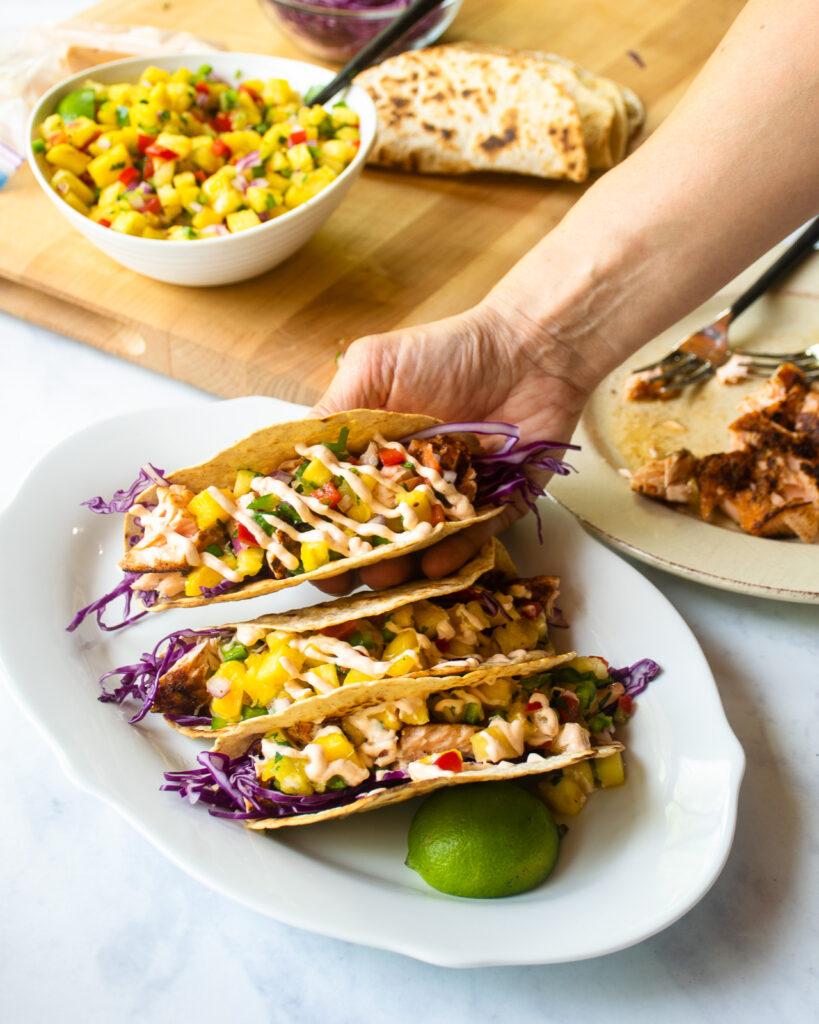 A hand holding a spicy salmon taco over a platter of others with ingredients around.