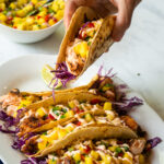 A hand lifting a spicy salmon taco from a plate of several tacos, with a bowl of pineapple salsa in the background.