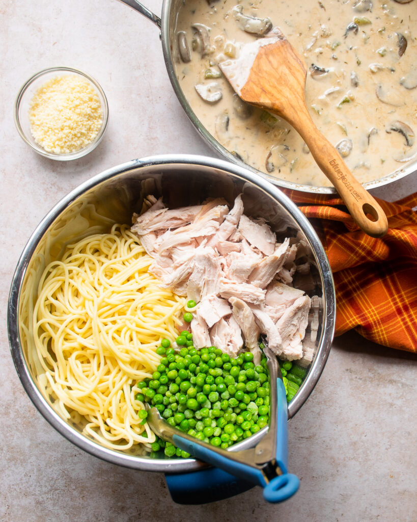 Looking down into a bowl with shredded turkey, cooked pasta and peas with a saute pan full of sauce and a bowl of parmesan cheese near by.