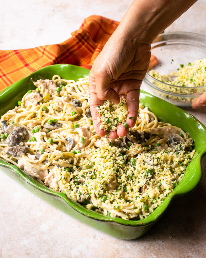 A hand sprinkling a breadcrumb topping onto a casserole of turkey tetrazzini.