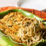 A spoon lifting turkey tetrazzini out of a green casserole dish with kitchen towels in orange and green around and a couple of white bowls in the background.