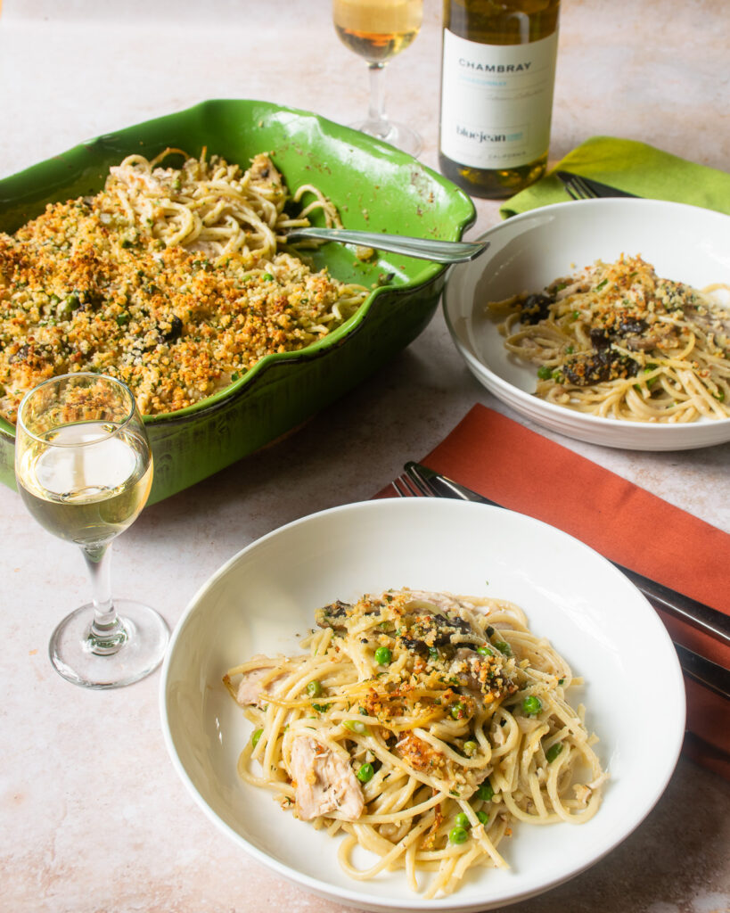 A serving of turkey tetrazzini in a white bowl with a glass of white wine and the rest of the casserole in the background.