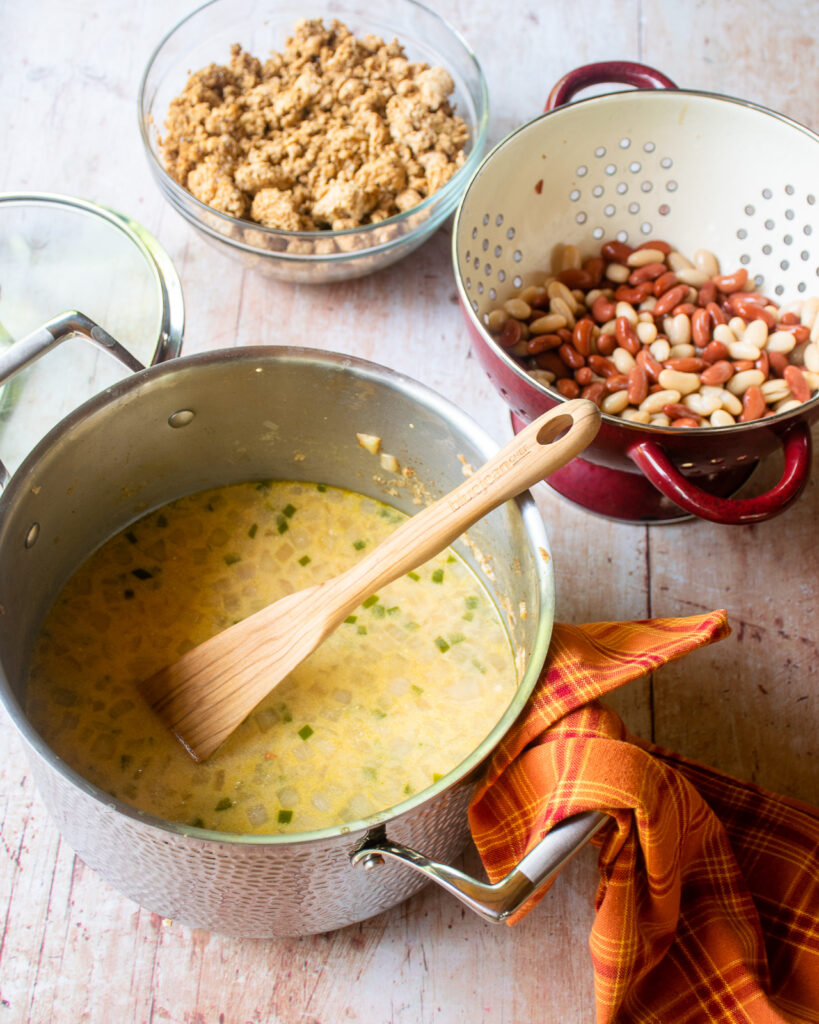 A soup pot of chili base with a wooden spoon sticking out of it next to a strainer of canned beans and a bowl of browned ground chicken.