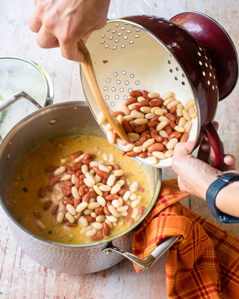 Hands adding beans to a soup pot full of white chicken chili.