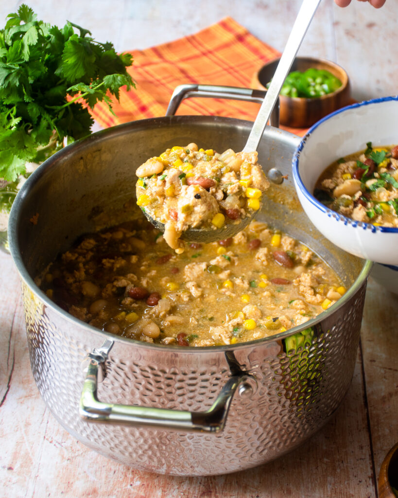 A ladle of white chicken chili being served into a small blue-rimmed white bowl.