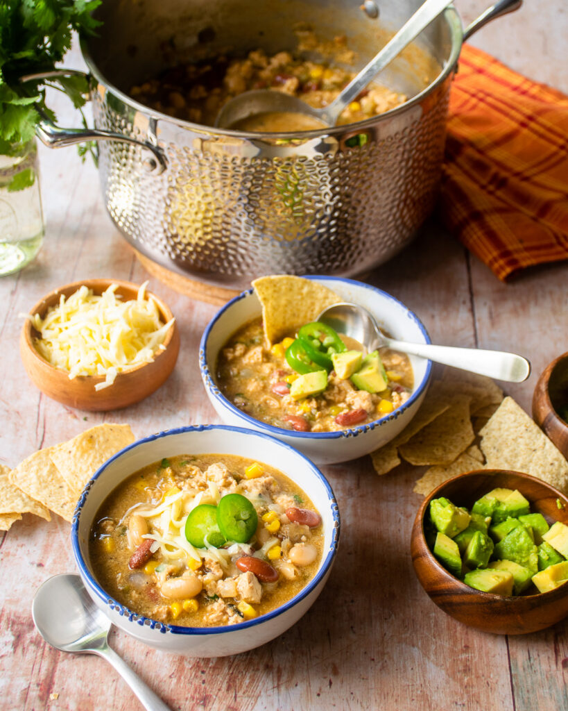 Two small bowls of white chicken chili next to a large stockpot of chili, some small bowls of jalapeño slices and grated cheese.