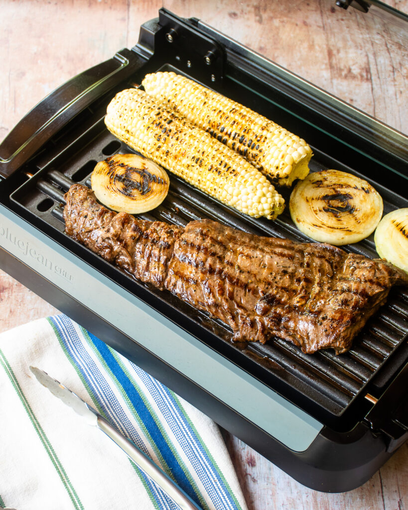 skirt steak grilling on an electric grill pan with corn on the cob and onion rings.